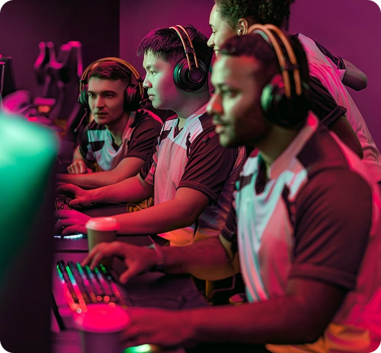 Vier geconcentreerde gamers met headsets en teamshirts spelen op computers in een schemerige gameroom met felroze en groene verlichting